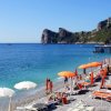 Spiaggia con lettini, ombrelloni arancioni e bianchi, vista mare e scogliera.