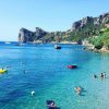 Mare limpido con bagnanti, scogliera sullo sfondo. Barche e canotti galleggiano sull'acqua.