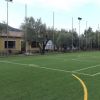 Un campo da calcio con erba sintetica circondato da alberi e un edificio giallo.