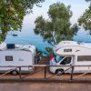 Due camper parcheggiati vicino a un mare calmo, circondati da alberi verdi.